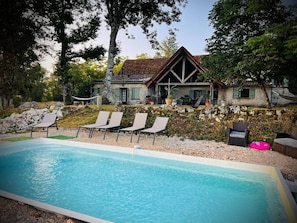 Ein Schwimmbecken mit Liegestühlen, ein Steinhaus mit geneigtem Dach und eine Steinmauer.