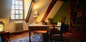 A room with a sloped ceiling, a table with a lamp, a chair, a bookshelf, and a radiator. - Under skies that arch from field to forest (Saint-Cernin-de-Labarde)