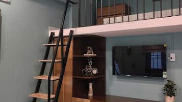A room with a wooden desk, a white chair, a TV, a bookshelf, and a staircase to an upper level.