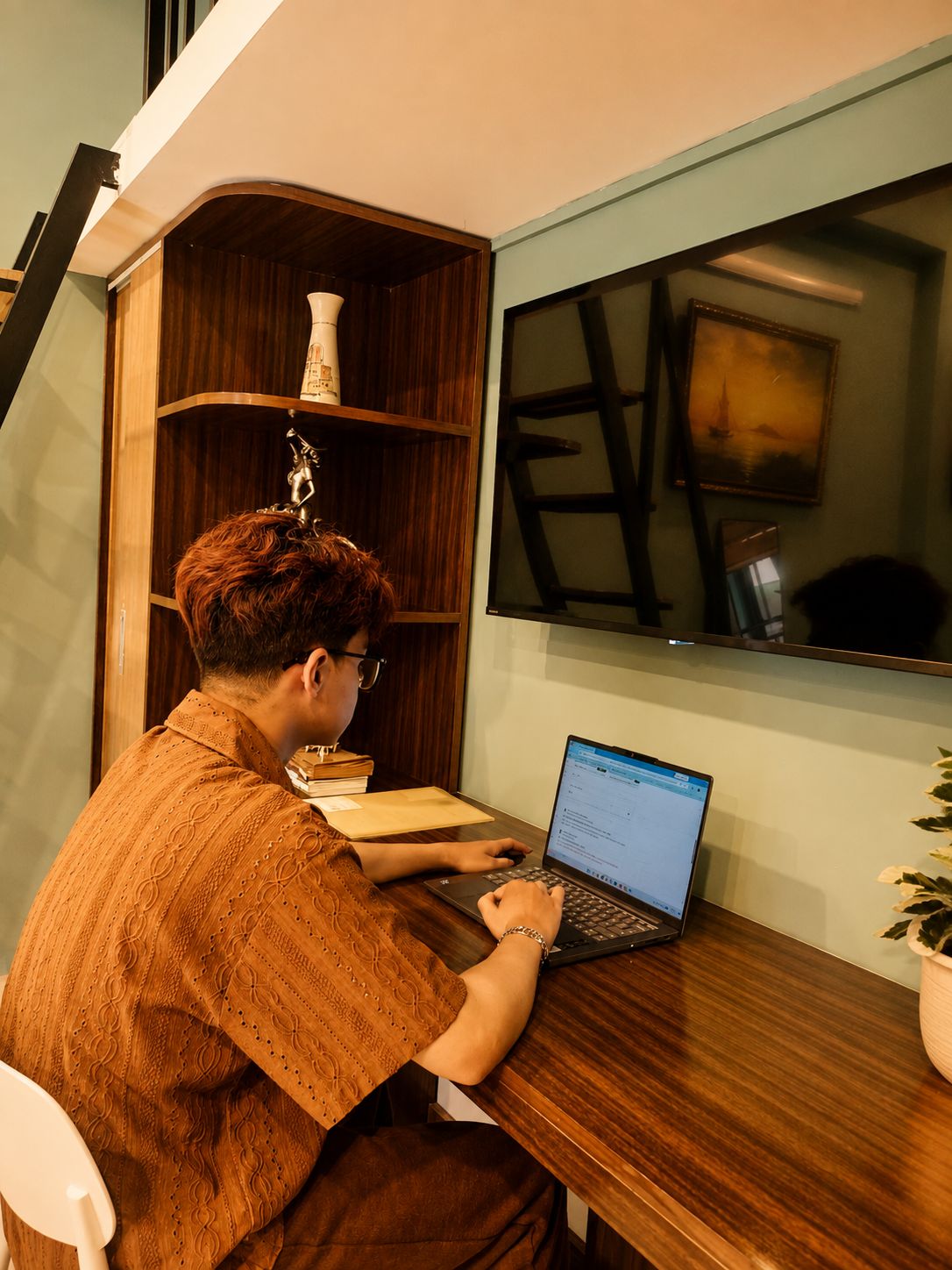 Une personne est assise à un bureau en bois, devant un ordinateur portable, dans une pièce où se trouve une bibliothèque en bois et un tableau encadré accroché au mur.