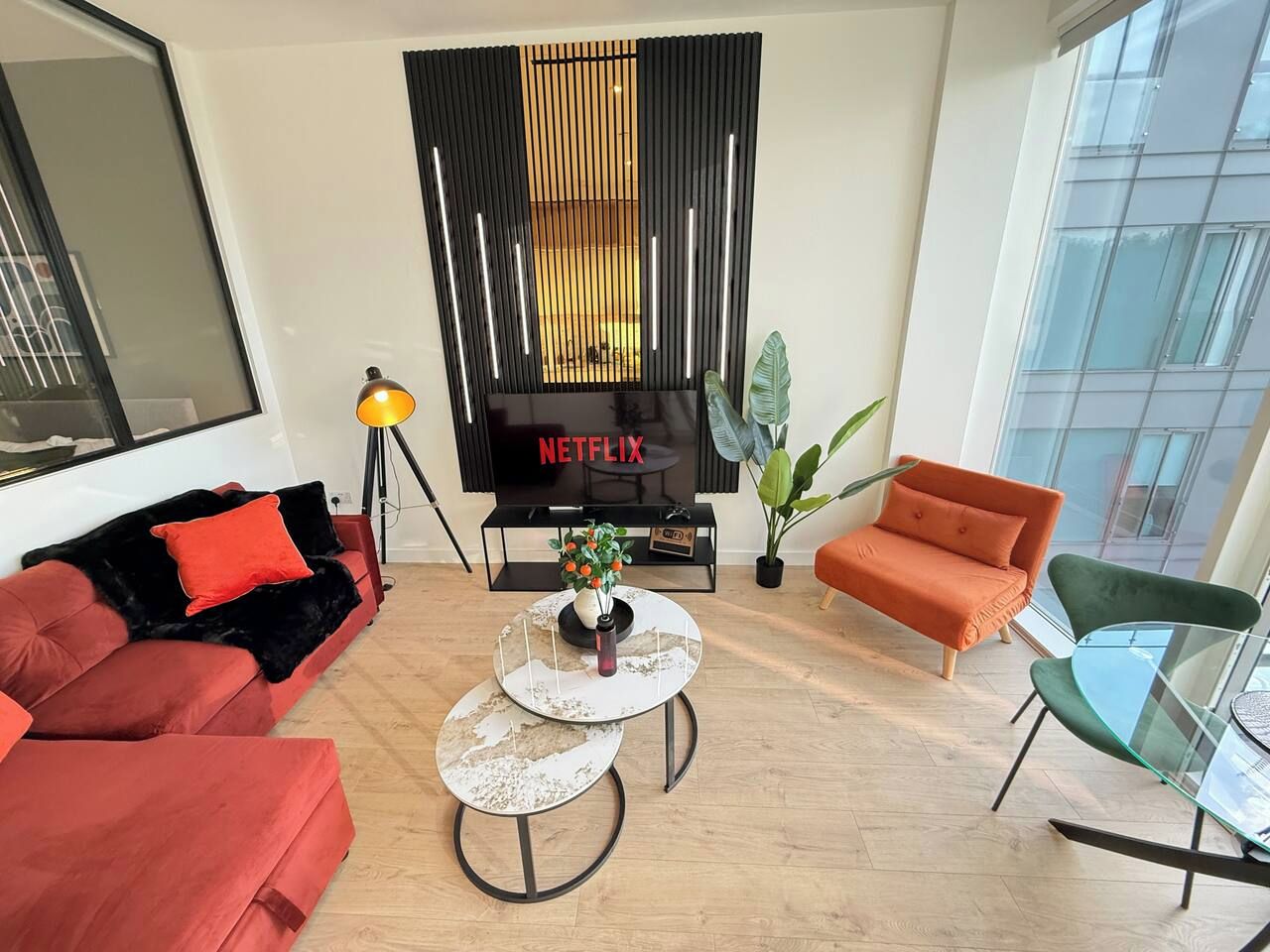 A modern living room with a flat-screen TV, red sofas, a glass coffee table, and a large window.