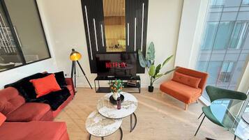 A modern living room with a flat-screen TV, red sofas, a glass coffee table, and a large window.