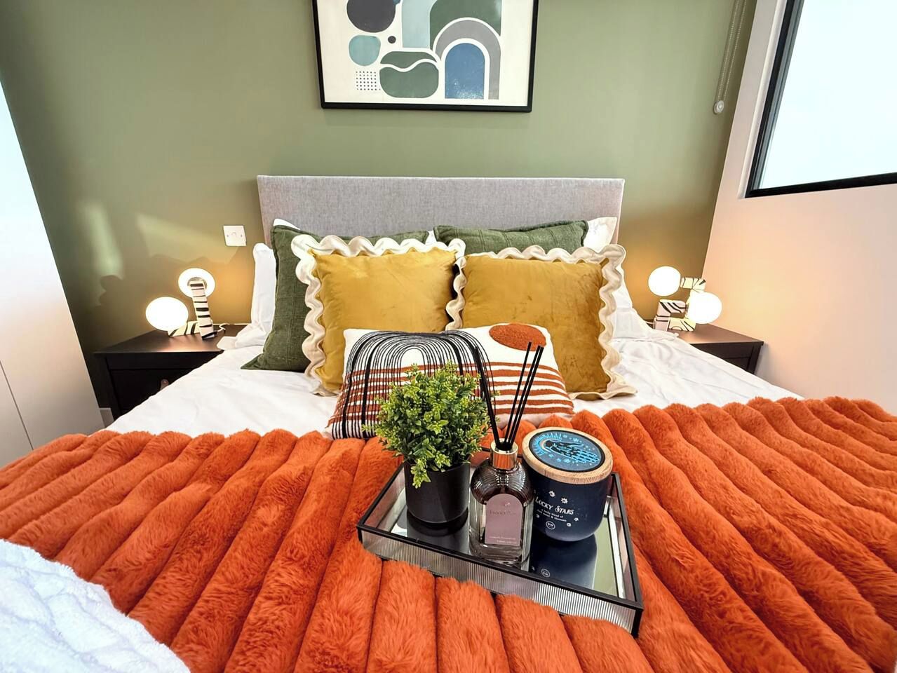 A neatly made bed with a fluffy orange blanket, a bedside table with a lamp, and a wall-mounted abstract art piece.