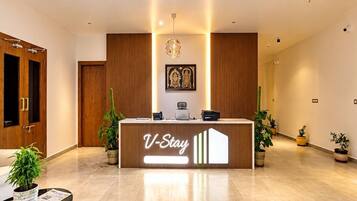 Una recepción con un cartel que dice 'V-Stay' en un moderno vestíbulo de hotel con paneles de madera, una araña y plantas en macetas.
