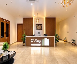 A reception desk with a sign reading 'V-Stay' in a modern hotel lobby with wooden paneling, a chandelier, and potted plants. - Classic V Stays (Chittoor)