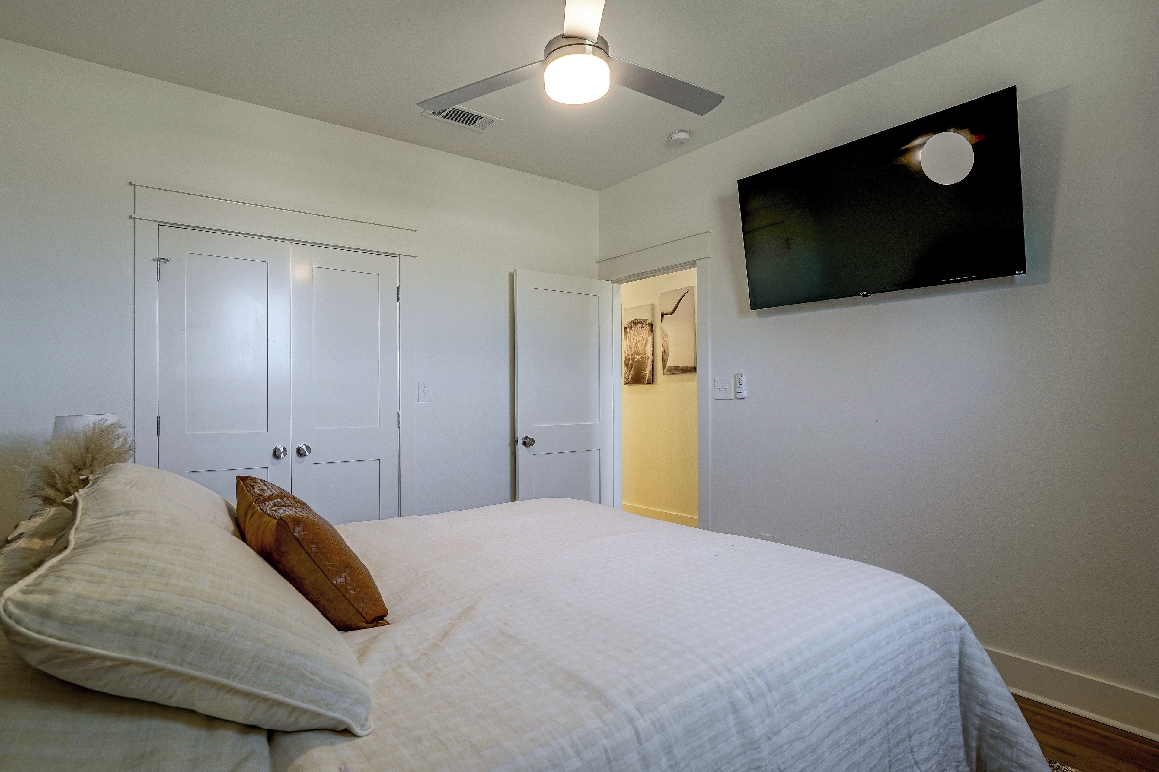 Une chambre à coucher comprenant un lit, un ventilateur de plafond, un téléviseur fixé au mur et une porte donnant sur un couloir.