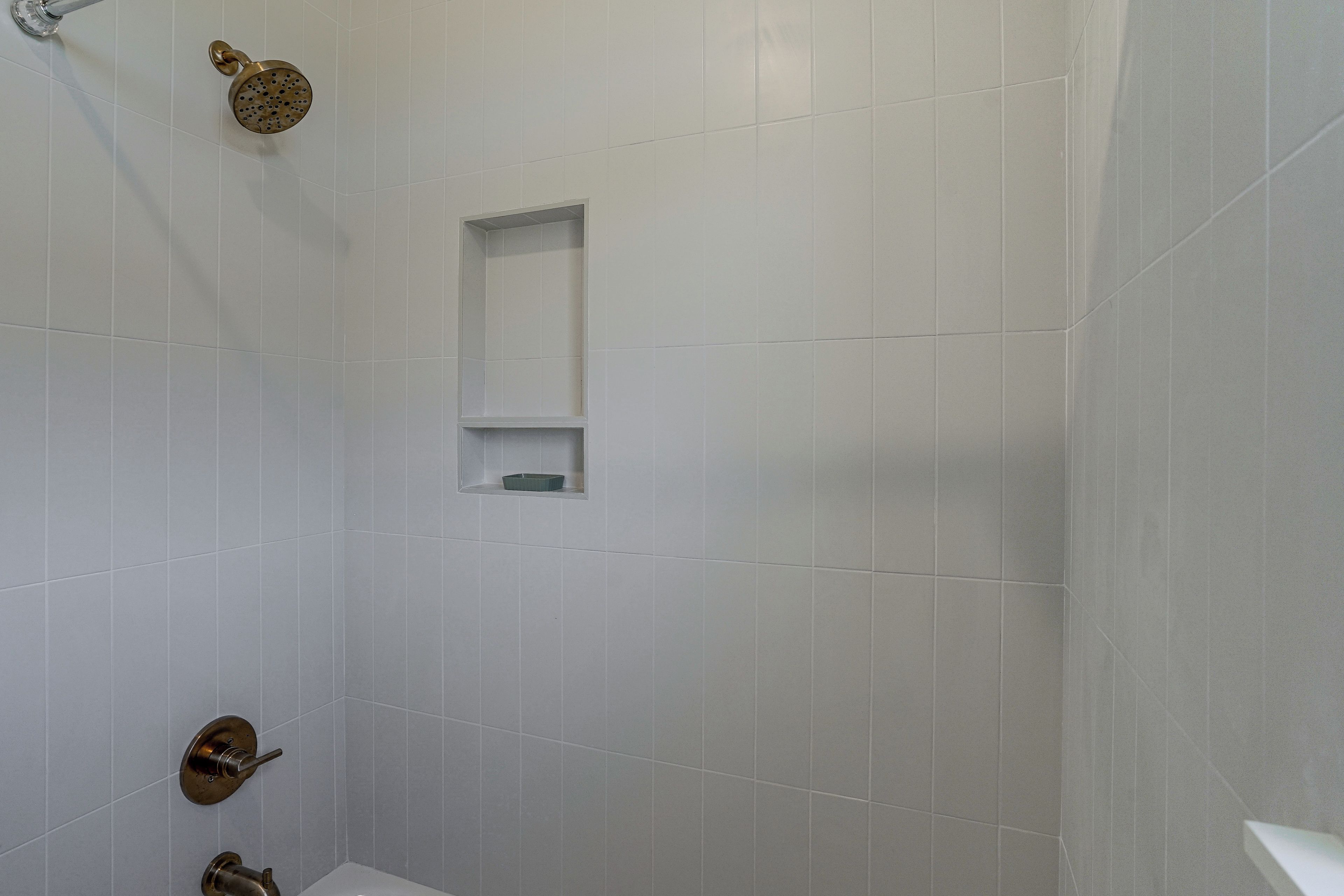 Une salle de bain aux murs recouverts de carreaux blancs, une douche dotée d’un pommeau doré et une petite étagère sur laquelle se trouve une bouteille.