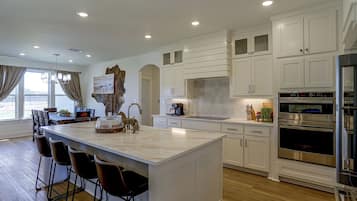 A modern kitchen with white cabinetry, a large island with bar stools, stainless steel appliances, and a view of the outdoors through a window.