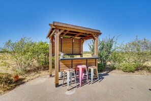 A wooden outdoor structure with a counter and stools, surrounded by desert vegetation. - 6-acre Barndominium Escape, 6 Mi to Lake Cleburne! (Cleburne)