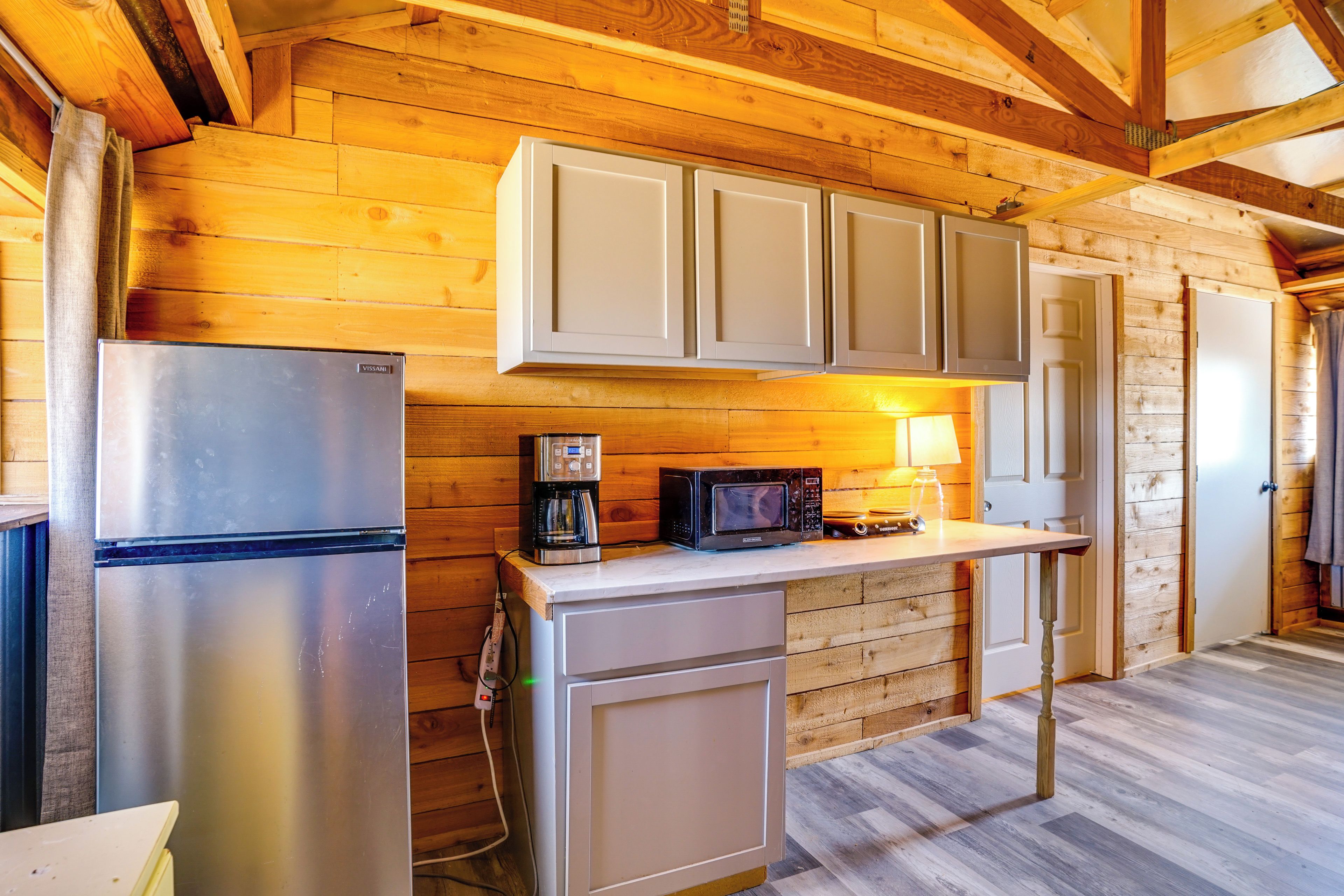 A compact kitchen with wooden walls, a stainless steel refrigerator, a microwave, and a coffee maker.