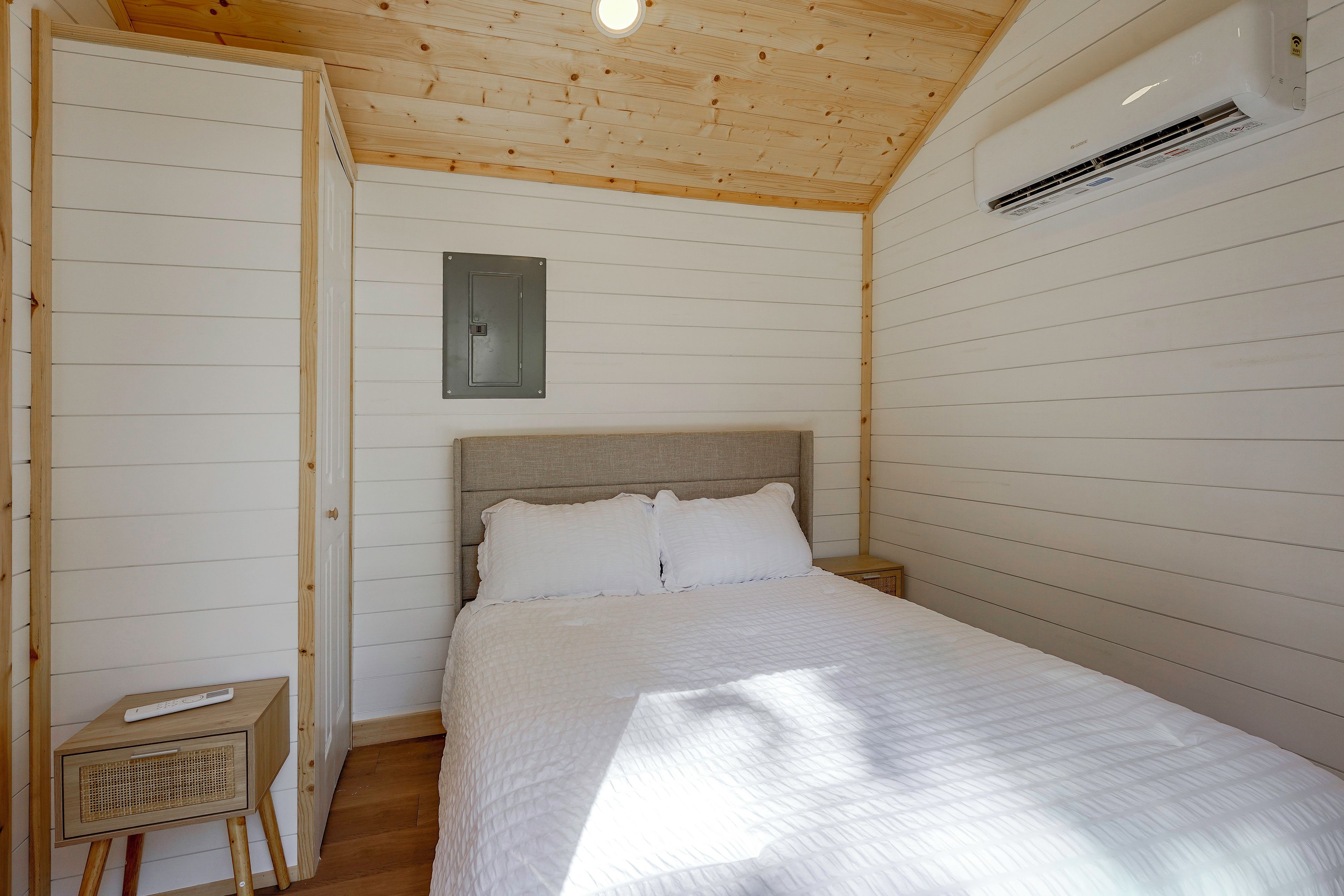 Un dormitorio con techo de madera, una cama con ropa de cama blanca, una mesita de noche y un aire acondicionado de pared.