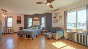 A bedroom with a bed, a sofa, a chair, a ceiling fan, and a window with a view of the street.