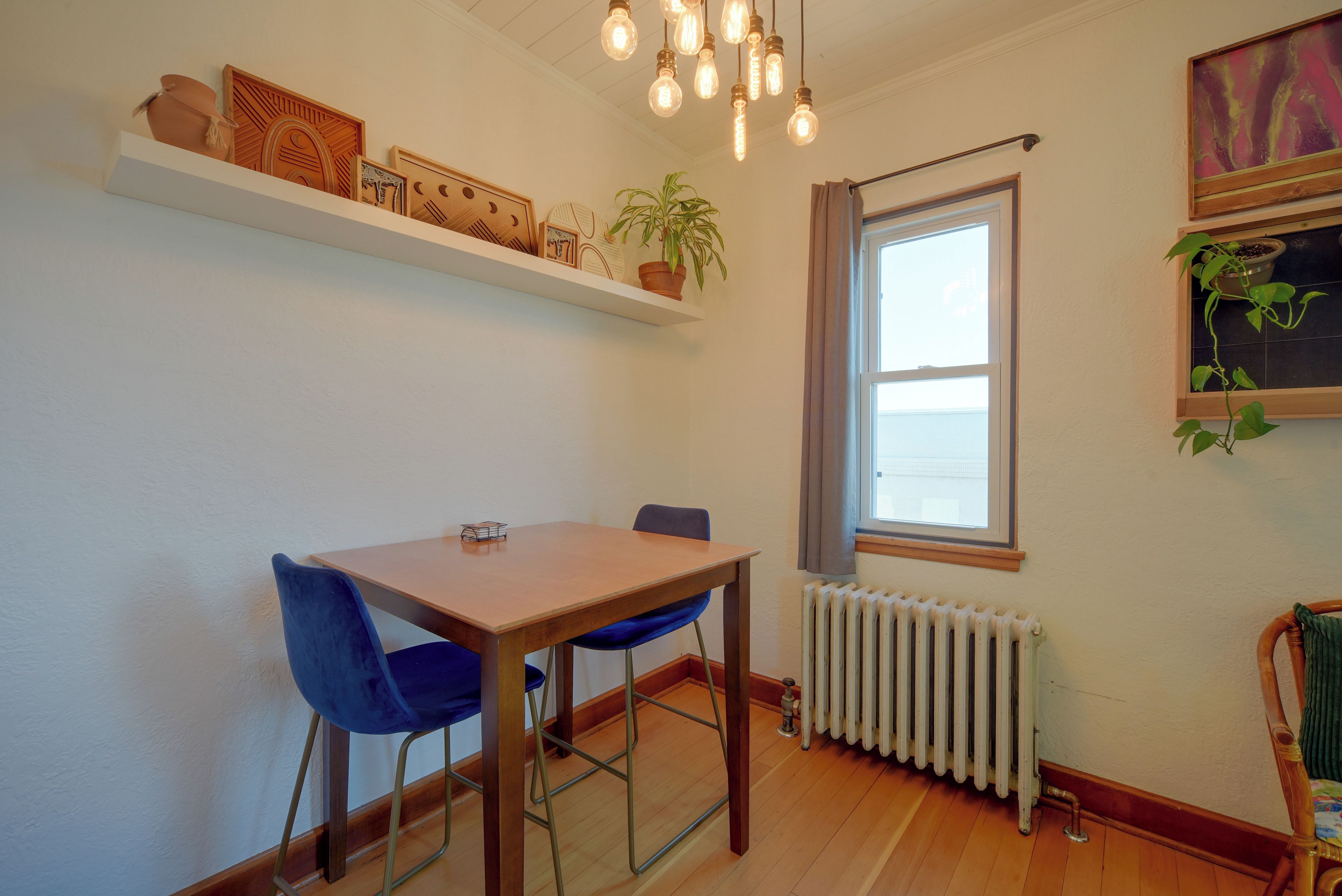 Un coin repas avec une table en bois et des chaises bleues, une plante en pot posée sur une étagère, un radiateur et une fenêtre avec des rideaux.