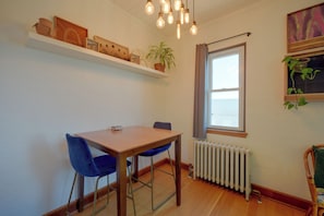 Un comedor con mesa de madera y sillas azules, una planta en maceta sobre una estantería, un radiador y una ventana con cortinas.
