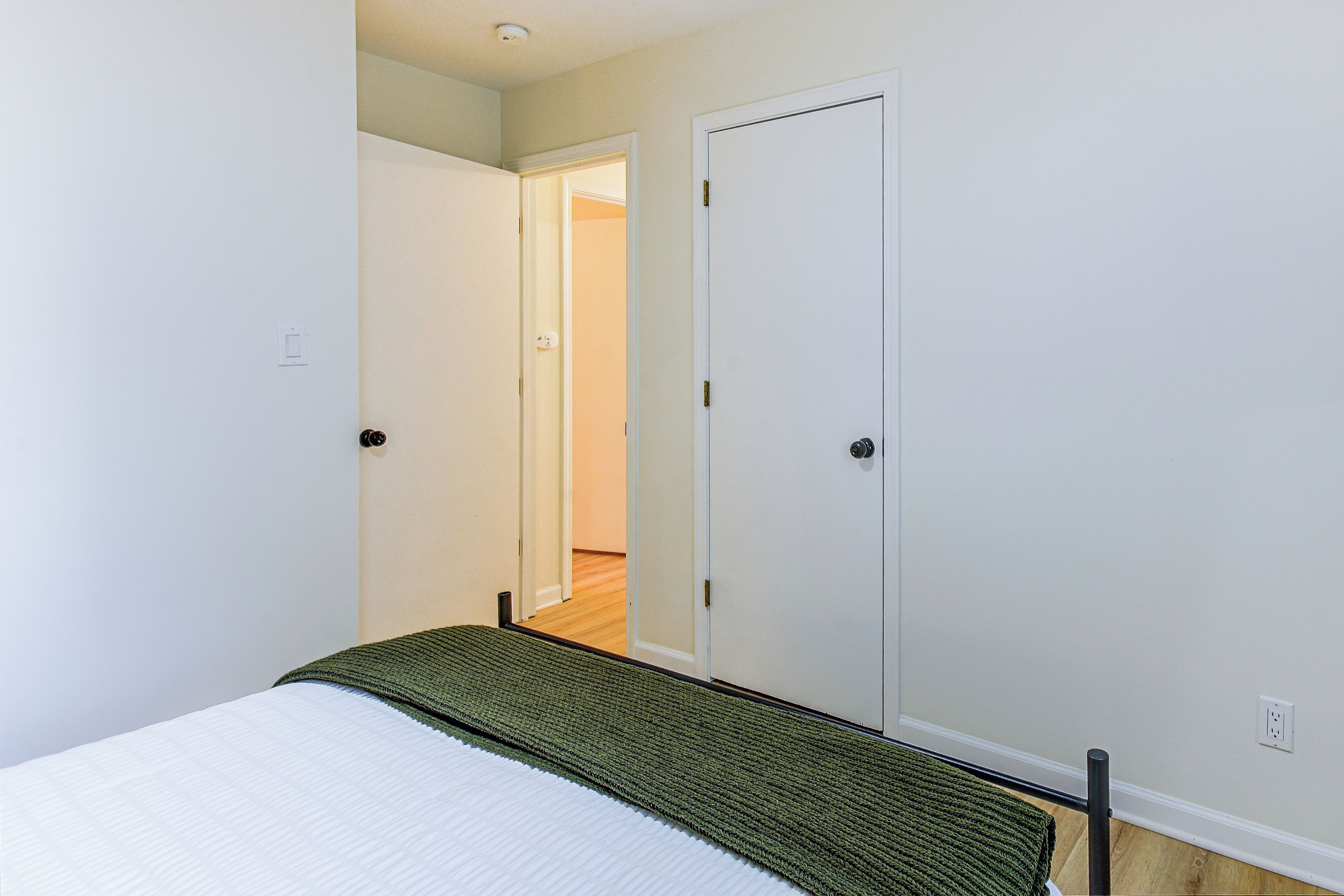 A narrow hallway with a bed at the end, a door on the right, and a light switch on the left wall.