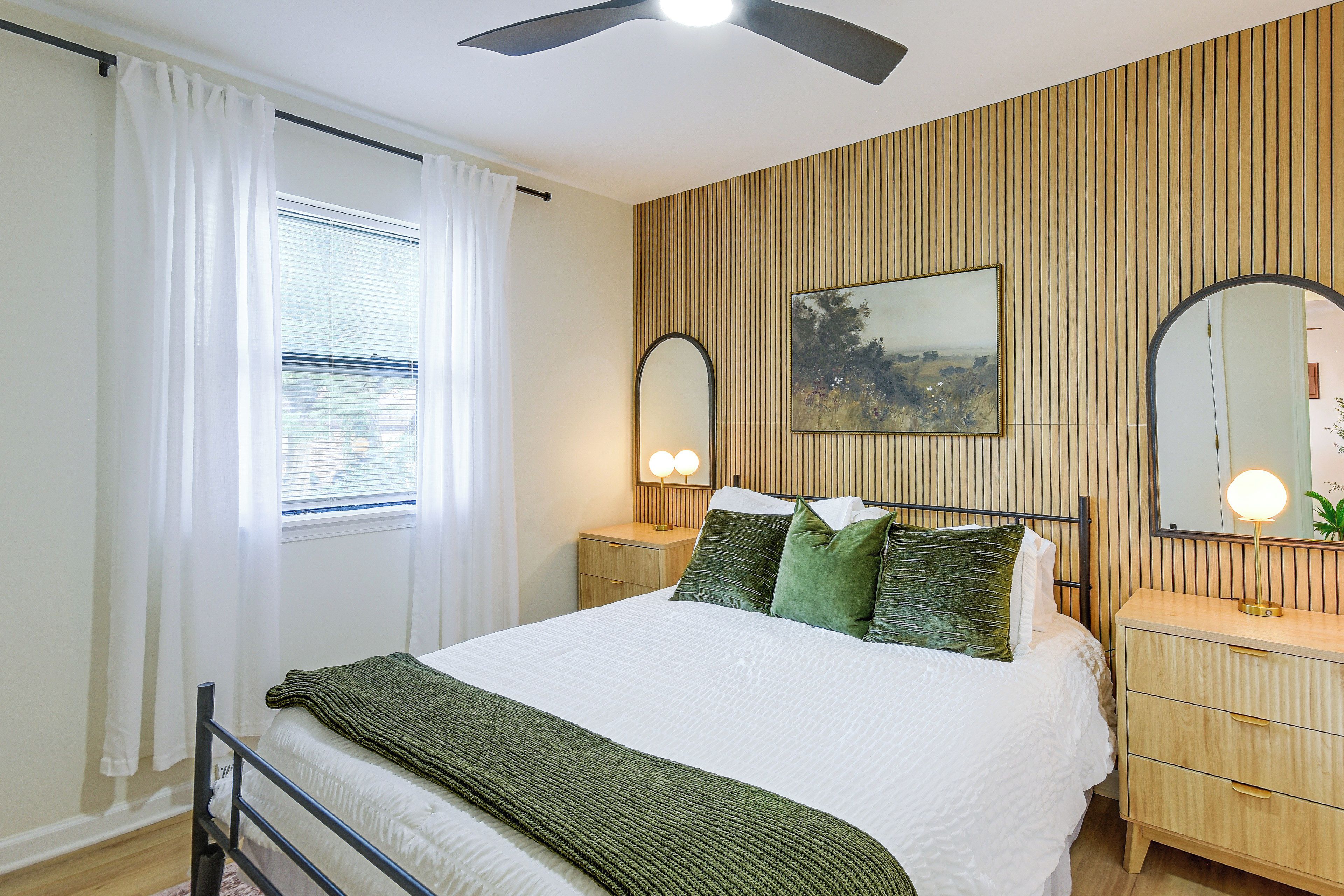 A bedroom with a bed, bedside tables, a mirror, a painting on the wall, and a window with curtains.
