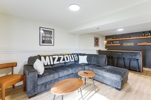 Un salon moderne avec un canapé d’angle gris, deux petites tables d’appoint en bois et un coin bar doté d’étagères.