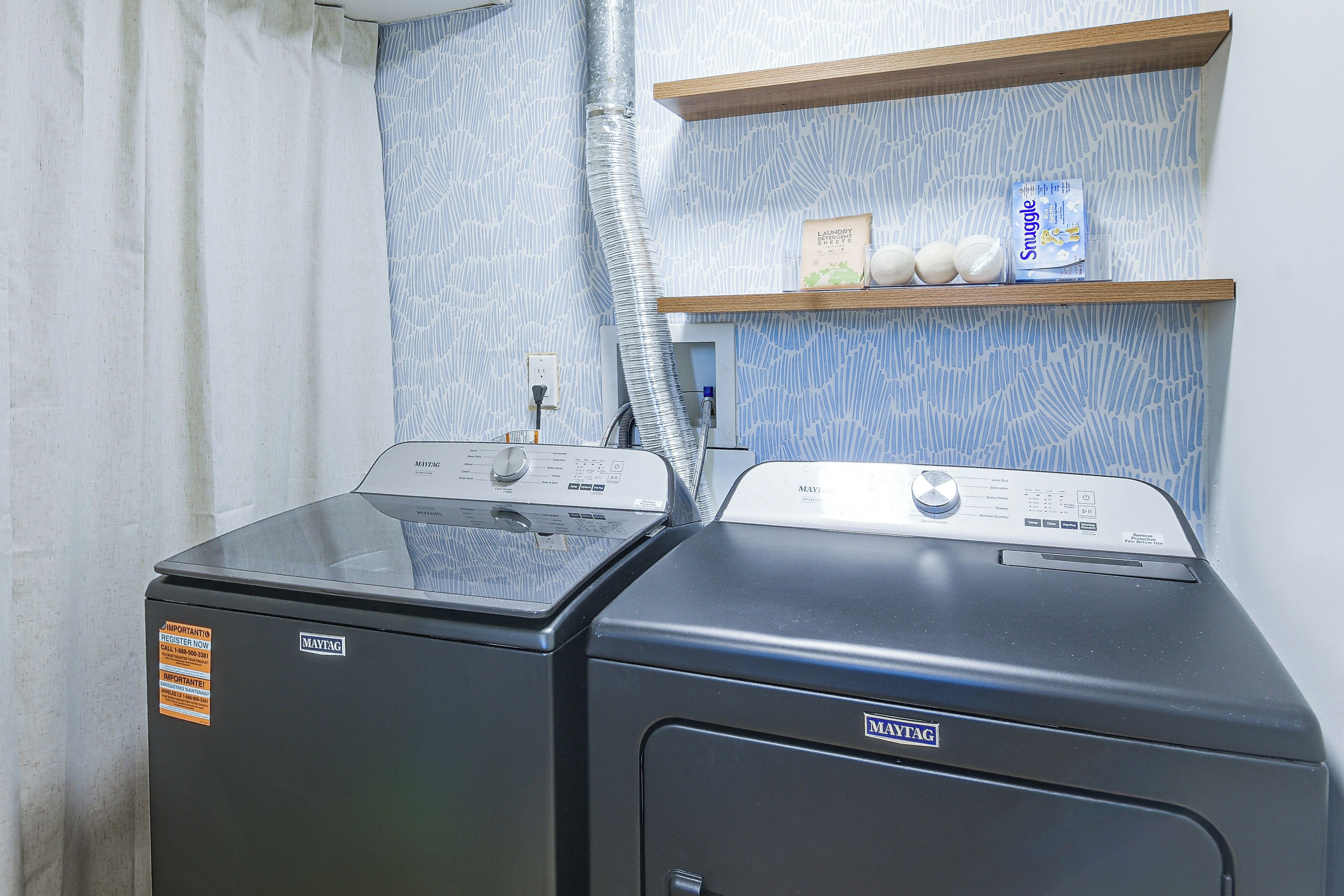 A laundry area with two Maytag washers and dryers, a shelf with laundry detergent, and a patterned wall.