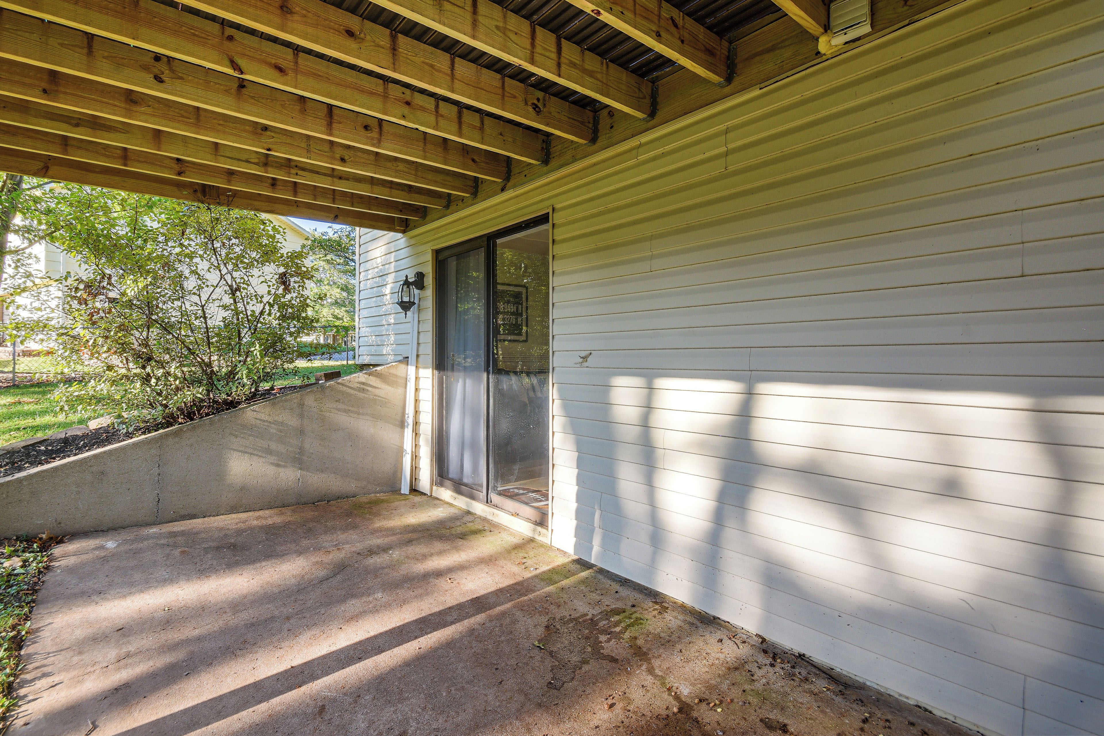 A covered porch with a concrete step leading to a door, wooden beams overhead, and a view of a residential area with trees and a house in the background.