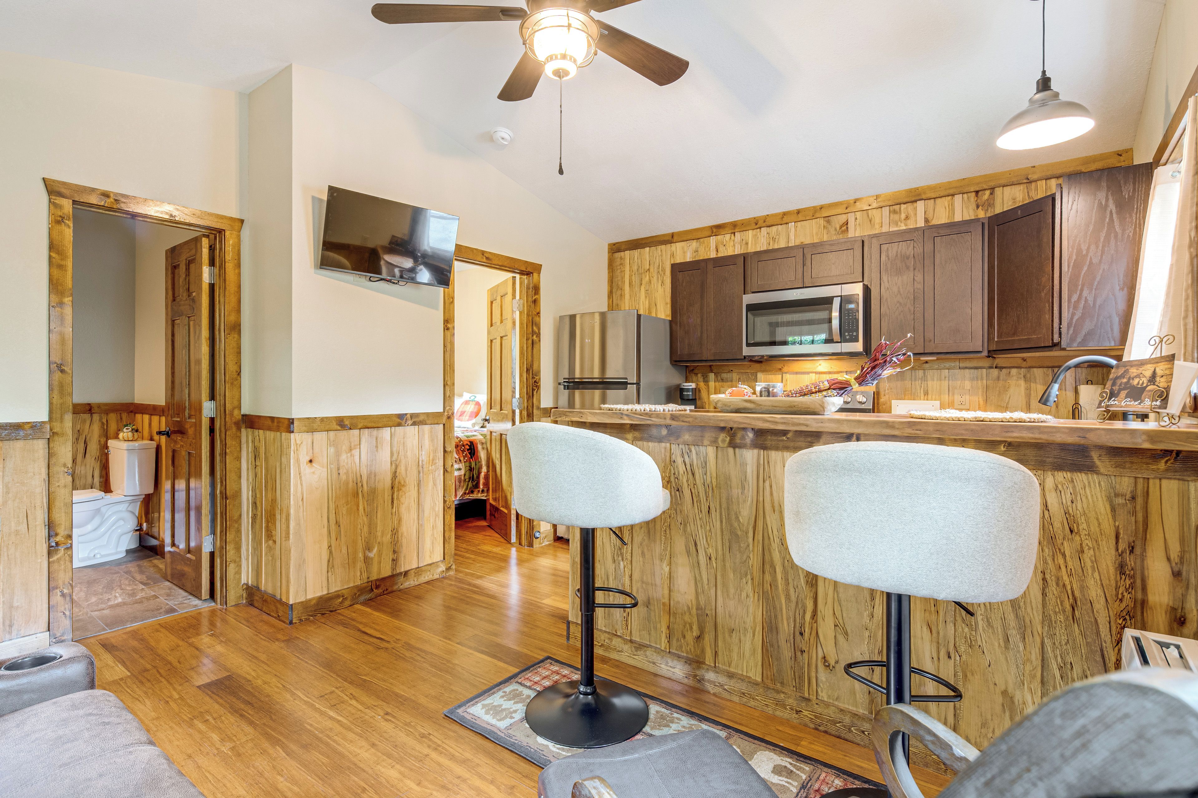 A kitchen with wooden cabinets, a microwave, and a refrigerator. There are two bar stools and a ceiling fan.