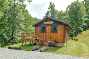 A wooden cabin labeled 'Deer Lodge' with a wooden deck, a stone foundation, and a gravel driveway. - Deer Lodge Cabin by the French Broad River (Brevard)