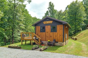 Uma cabana de madeira com a placa 'Deer Lodge', um deck de madeira, fundação de pedra e entrada por um caminho de cascalho.