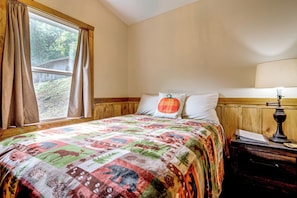 A bedroom with a wooden headboard, a quilted bedspread, a nightstand with a lamp, and a window with curtains. - Deer Lodge Cabin by the French Broad River (Brevard)