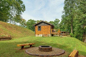 A wooden cabin with a stone fire pit surrounded by a gravel circle, set on a grassy hill with a wooden deck and a bench. - Deer Lodge Cabin by the French Broad River (Brevard)