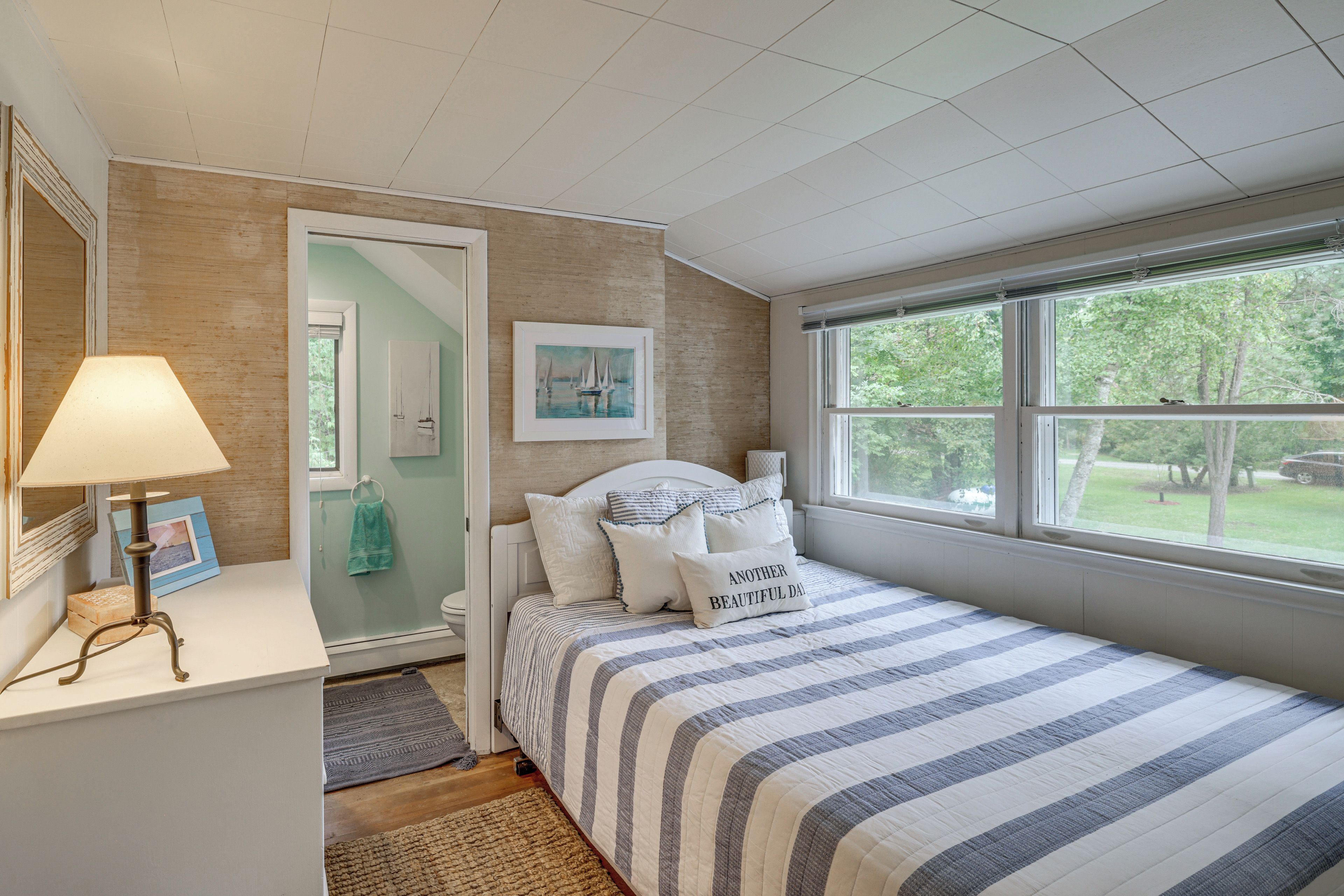 A bedroom with a bed, a bedside table with a lamp, a window with a view of trees, and a door leading to another room.