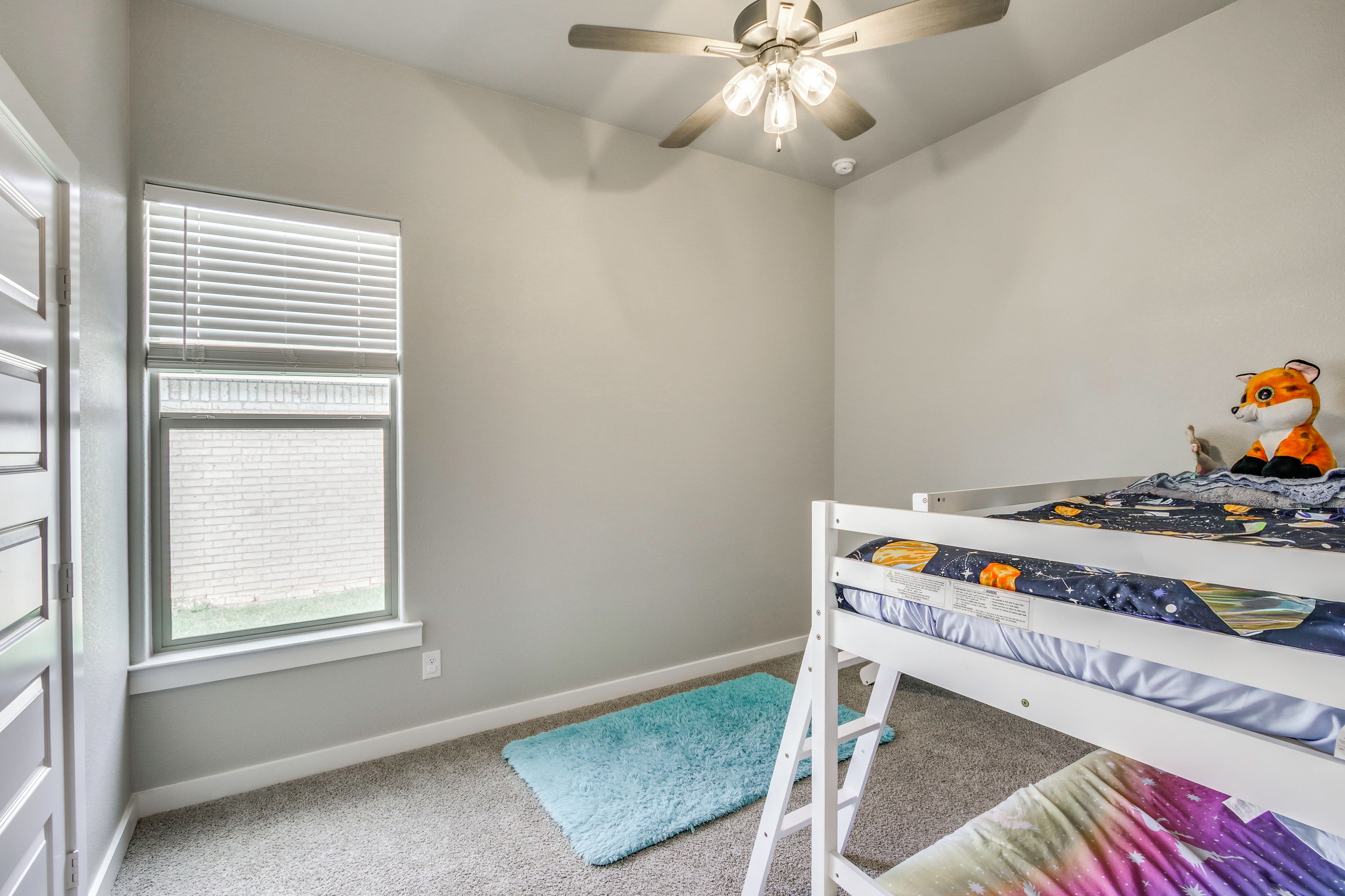 A child's bedroom with a bunk bed, a ceiling fan, a window with blinds, and a plush toy on the bed.