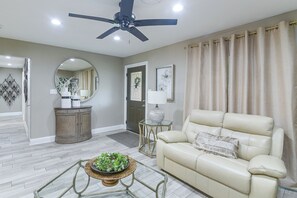 A living room with a ceiling fan, a beige sofa, a coffee table with a plant, and a door with a mirror and artwork.
