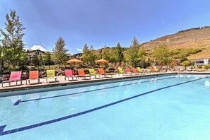 Ein Schwimmbecken mit mehreren Bahnen, umgeben von Liegestühlen und Sonnenschirmen, vor einer Kulisse aus Bäumen und einer hügeligen Landschaft.