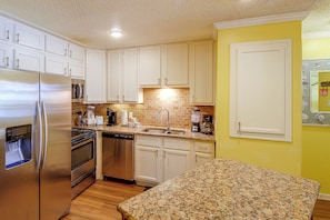 A kitchen with white cabinets, stainless steel appliances, a granite countertop, and a yellow accent wall. - Unwind Poolside, Walk to Shore! Port Aransas Condo (Port Aransas)