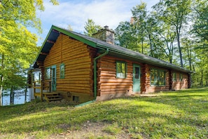 A log cabin with a porch and a chimney, surrounded by trees and a grassy area. - North Woods Log House w/ Exceptional Privacy! (Stone Lake)