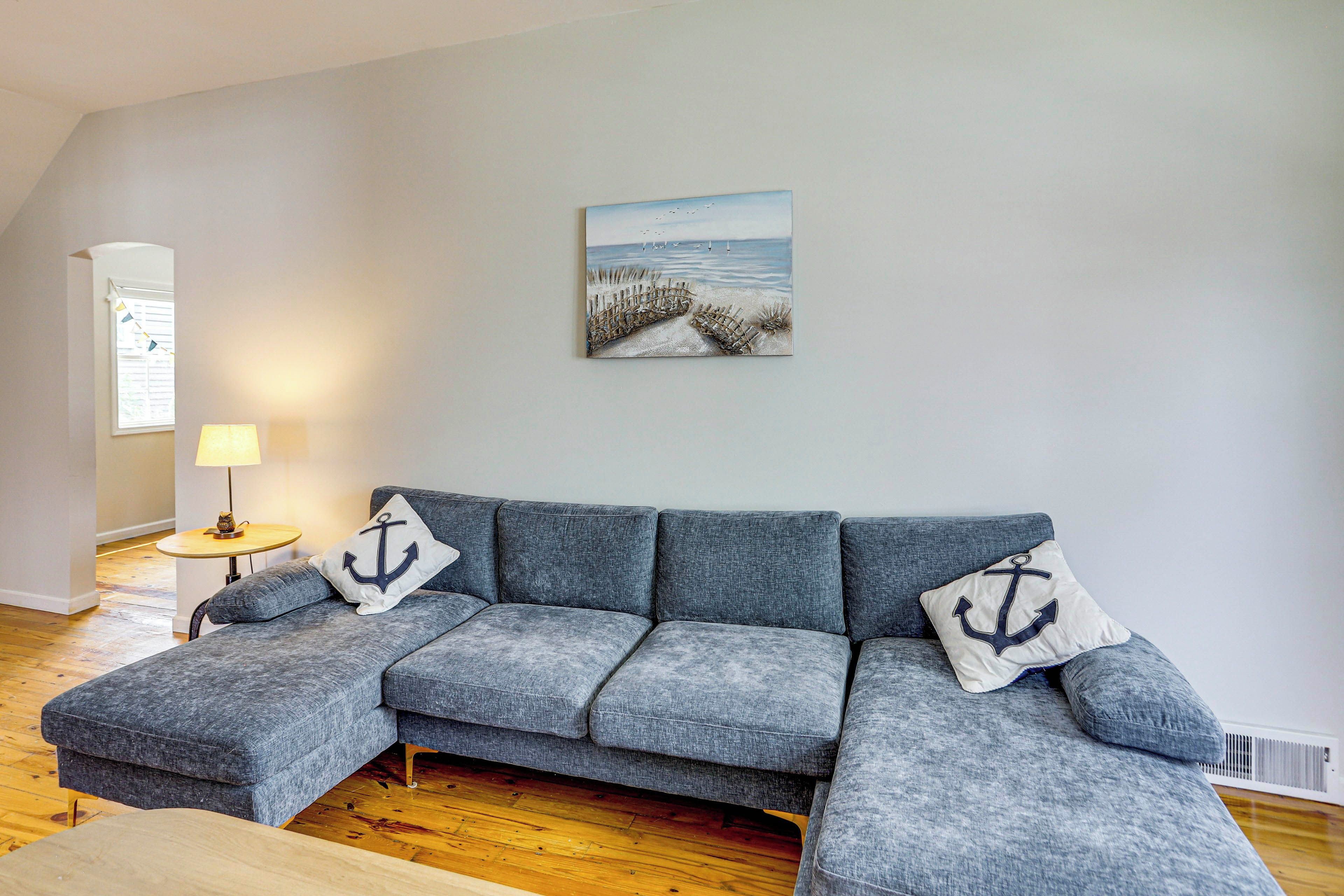 A living room with a grey sectional sofa, two anchor pillows, a painting of a beach scene, a lamp, and a window with blinds.