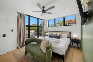 Ein modernes Schlafzimmer mit einem großen Bett, einem grünen Sessel, einem Deckenventilator und Blick ins Freie durch große Fenster.