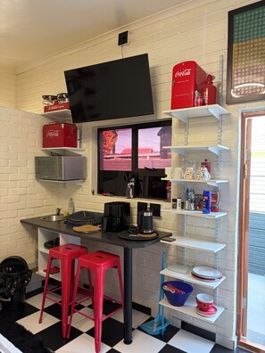 A compact kitchen with a flat-screen TV, red Coca-Cola crates, a coffee maker, and a sink. - The Cola Cabana (Cape Town)