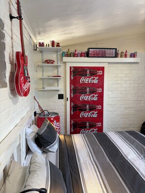 Una habitación con una cama, una guitarra, estantes con botellas y una puerta con el diseño de Coca-Cola.