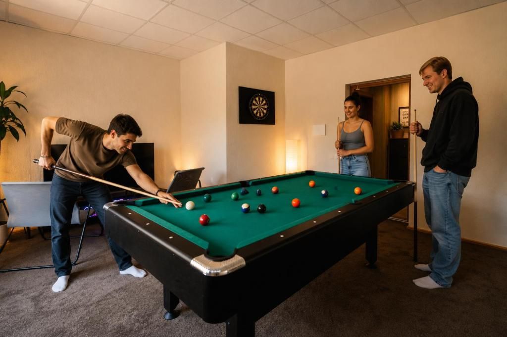 Ein Billardtisch in einem Raum mit Dartscheibe an der Wand, einer Pflanze und einer Tür zu einem anderen Raum.