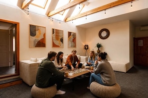 A group of people playing board games in a room with a sloped ceiling, abstract art on the walls, and a clock. - Hang the Hat (Valmiera)