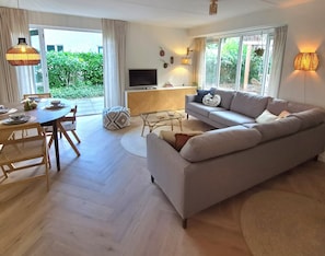 A modern living room with a sofa, dining table, and chairs. There is a television on a stand and a window with curtains. - Gemütliches Ferienhaus für 2-6 Personen mit privater Hottub in schönen Lochem (Lochem)