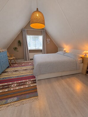 Une chambre à coucher avec un lit, des lampes de chevet, un luminaire suspendu, une commode, une chaise, une fenêtre avec des rideaux et un tapis à motifs.