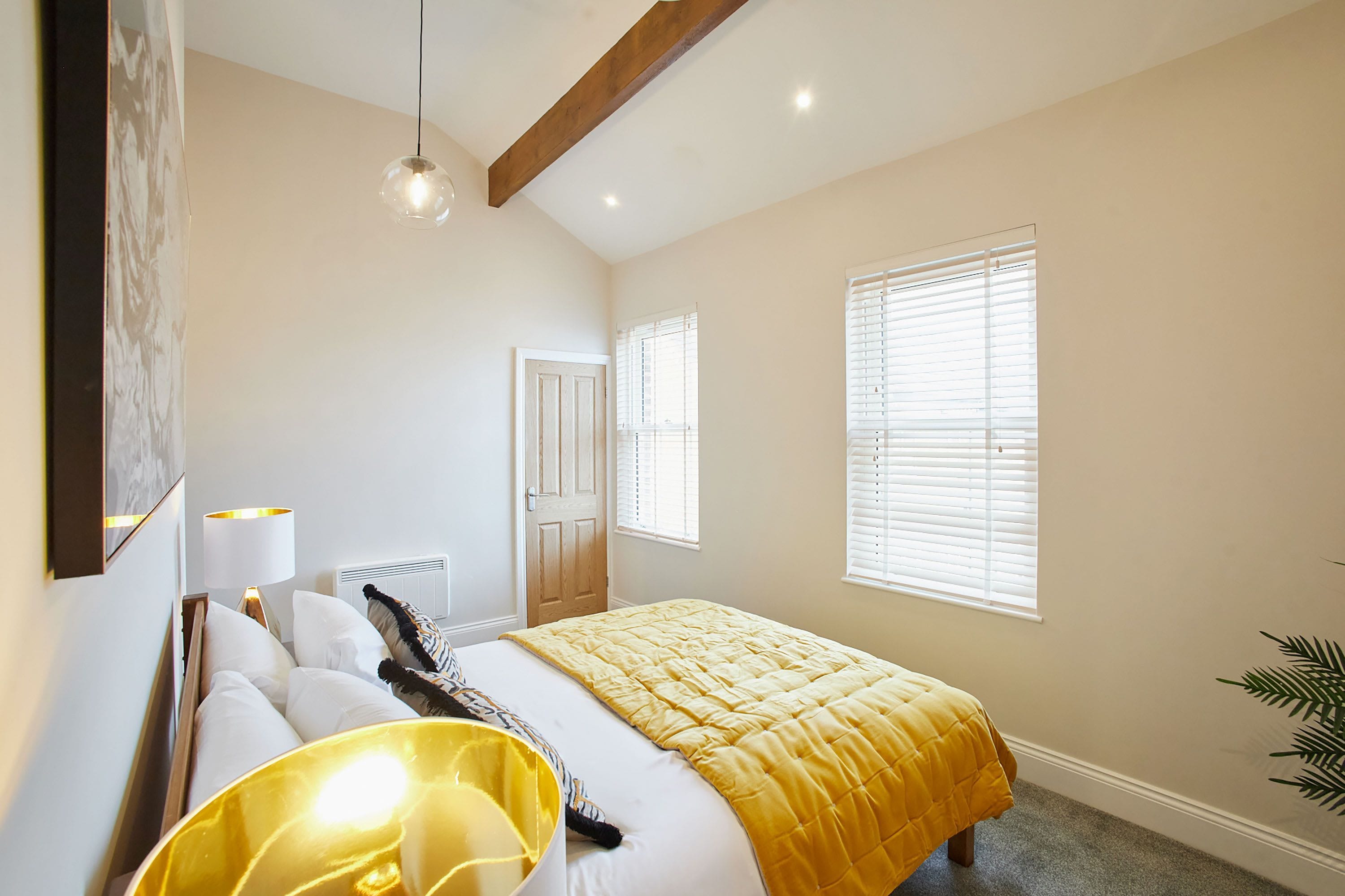 A bedroom with a bed, a bedside lamp, a window with blinds, a radiator, and a plant.