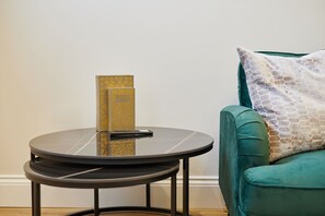 A round table with a remote control and a book, a teal sofa with a patterned pillow, and a plain wall. - Host Stay - Temperance Mews (Saltburn-by-the-Sea)