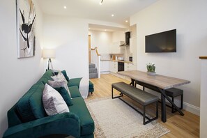 A modern living room with a green sofa, a wooden dining table, and a flat-screen TV mounted on the wall. - Host Stay - Temperance Mews (Saltburn-by-the-Sea)
