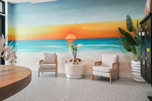Ein Zimmer mit Wandgemälde vom Strand, zwei Korbstühle, einem runden Couchtisch und einer Topfpflanze.