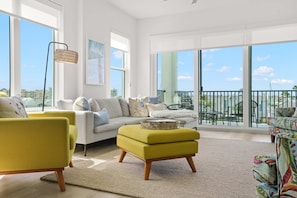 Ein geräumiges Wohnzimmer mit einer Couch, Hocker und Sesseln, großen Fenstern und einem Balkon mit Gartenmöbeln.