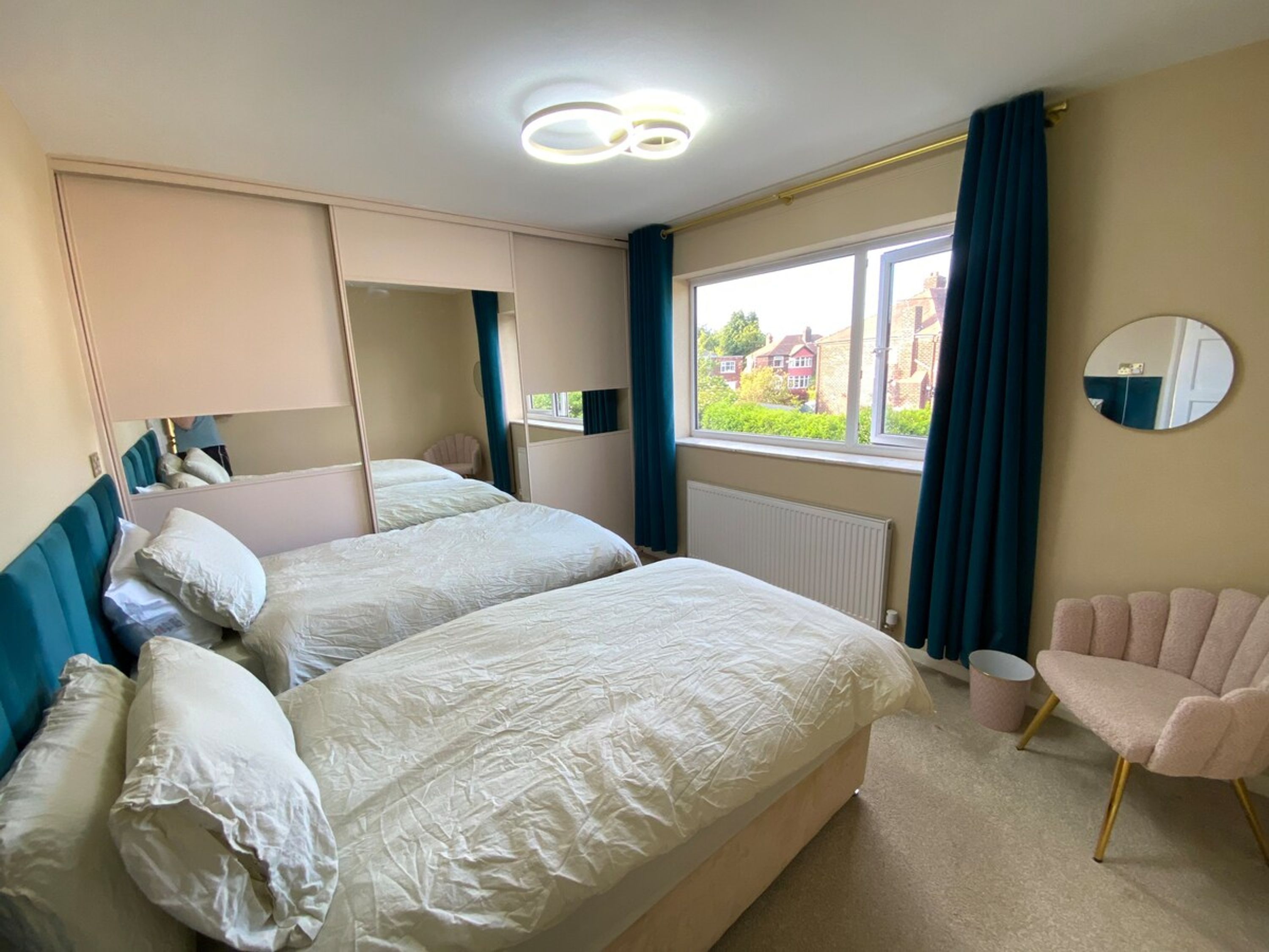 A hotel room with two beds, a chair, a large mirror, and a window with a view of houses.