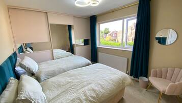 A hotel room with two beds, a chair, a large mirror, and a window with a view of houses.