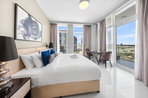 A hotel room with a large bed, a desk with a lamp, a chair, and a view of the cityscape through large windows. - Stayshort - Cozy Studio in Damac Hills 1 for 2 With Pool Access (Dubai)